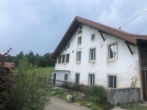 A two-story building with a sloped roof, multiple windows, and a small garden area with a metal fence. - Appartement et B&B - Ferme O'Badu, (Les Breuleux), by Interhome (Les Breuleux)