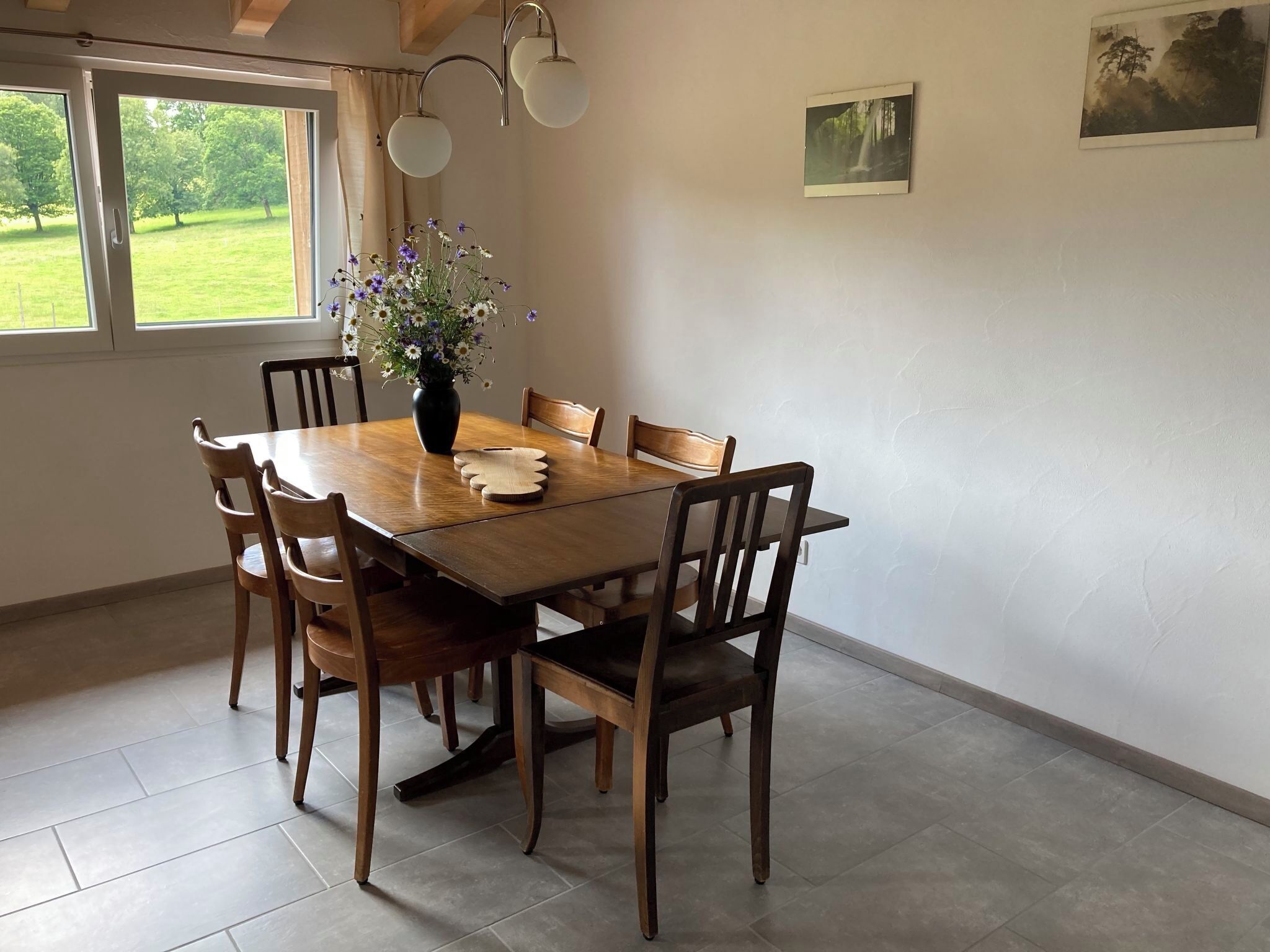 A dining area with a wooden table and chairs, a vase with flowers, and a view of greenery outside.