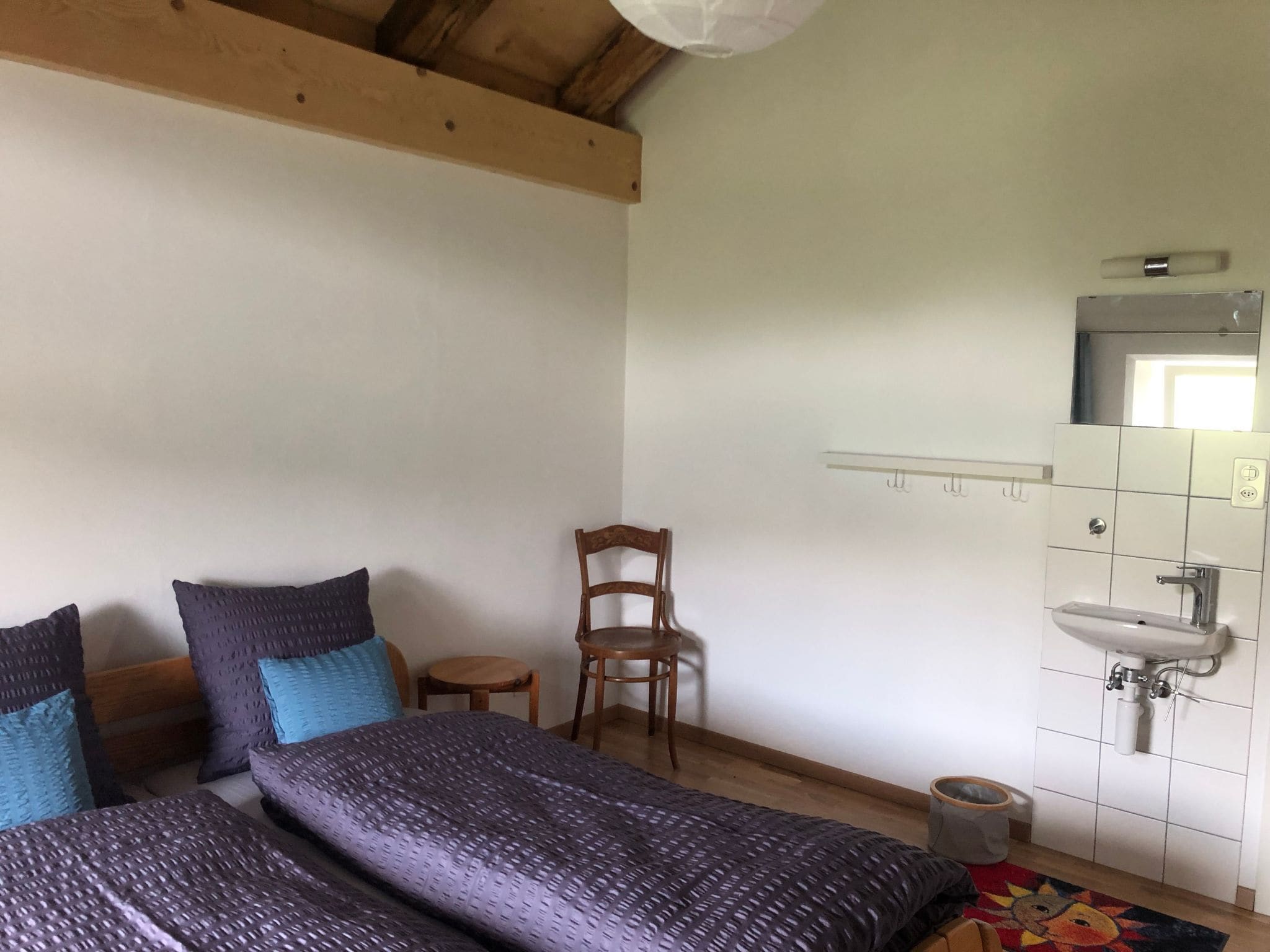 Quarto com duas camas, uma cadeira de madeira, uma mesinha, uma pia e um espelho.