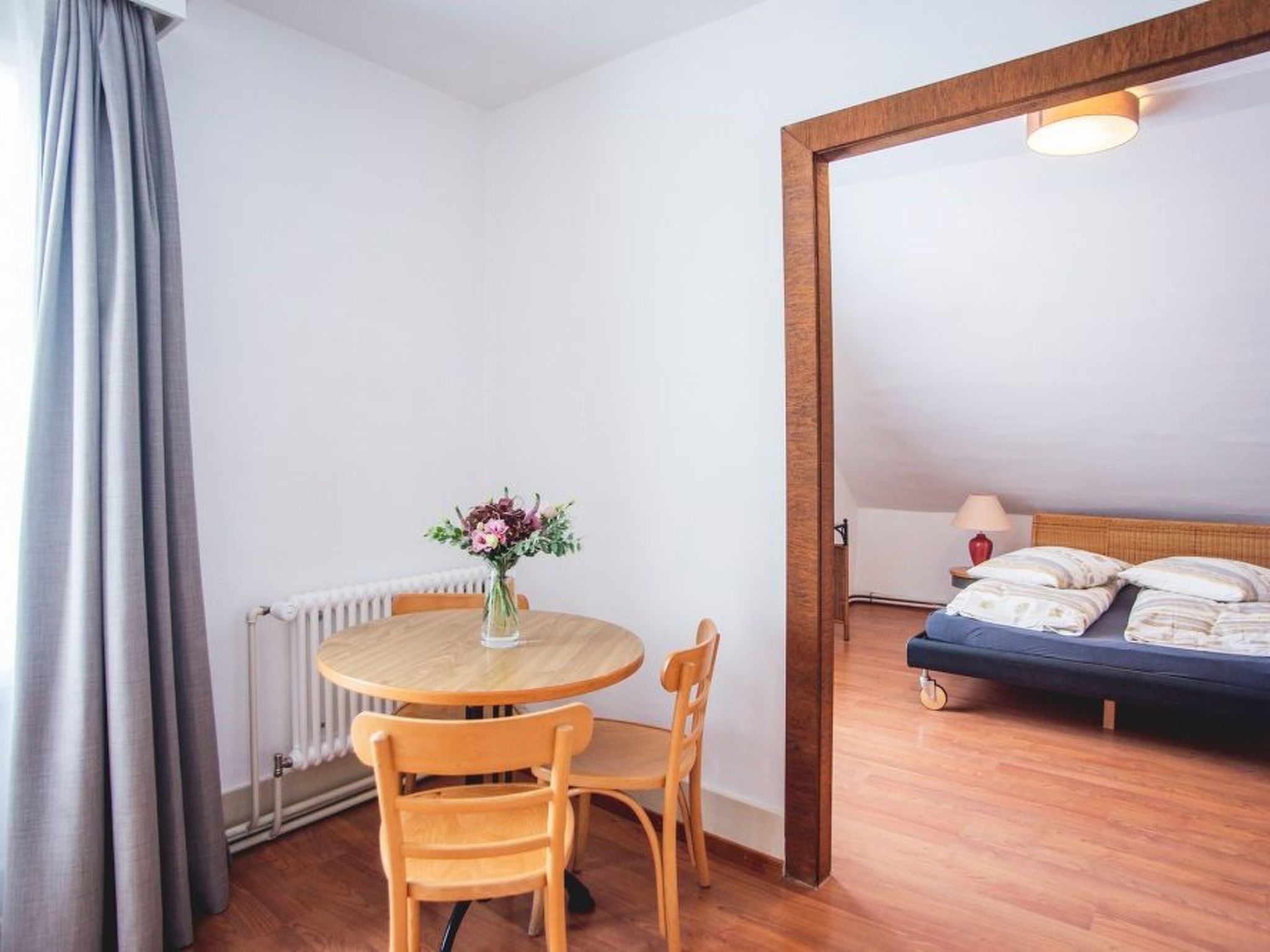 Ein Zimmer mit einem runden Holztisch, zwei Stühlen, einem Bett mit Kopfteil, einem Heizkörper und einem Fenster mit Vorhängen.