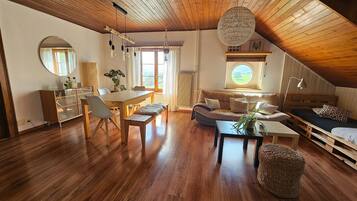 A spacious room with a wooden ceiling, a dining area, a sofa, a window with a view, and a hanging light fixture.