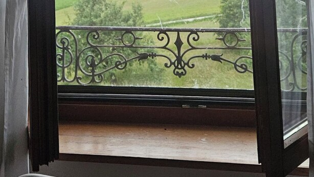 Udsigt gennem et vindue med en balkon, der viser et landskab med marker og fjerne bjerge under en skyet himmel.
