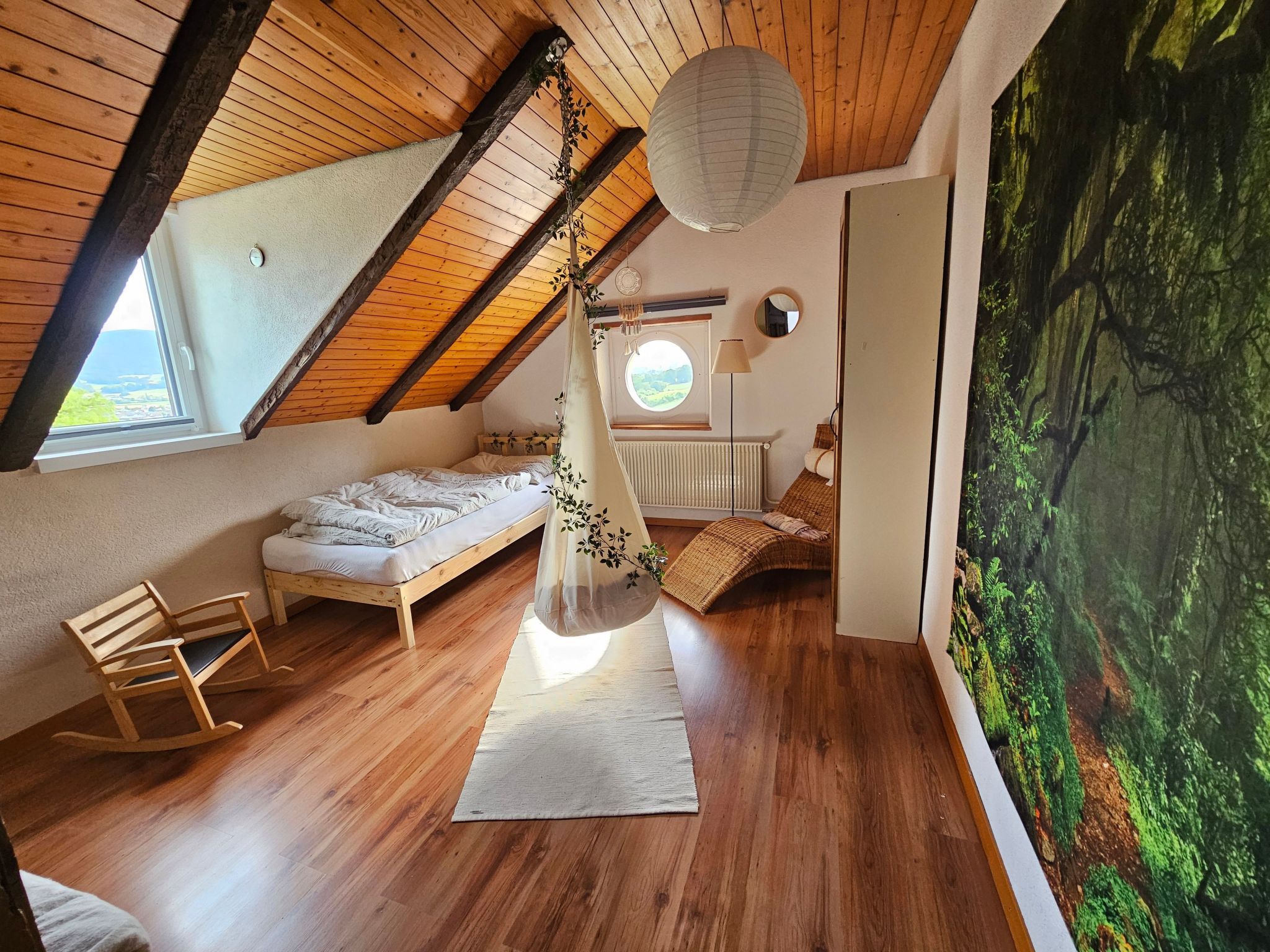 A cozy attic bedroom with a sloped ceiling, a bed, a rocking chair, and a large window.