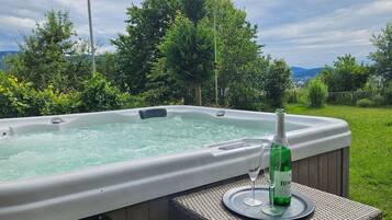 A hot tub with a bottle of wine and glasses on a table, surrounded by greenery and a view of the landscape.