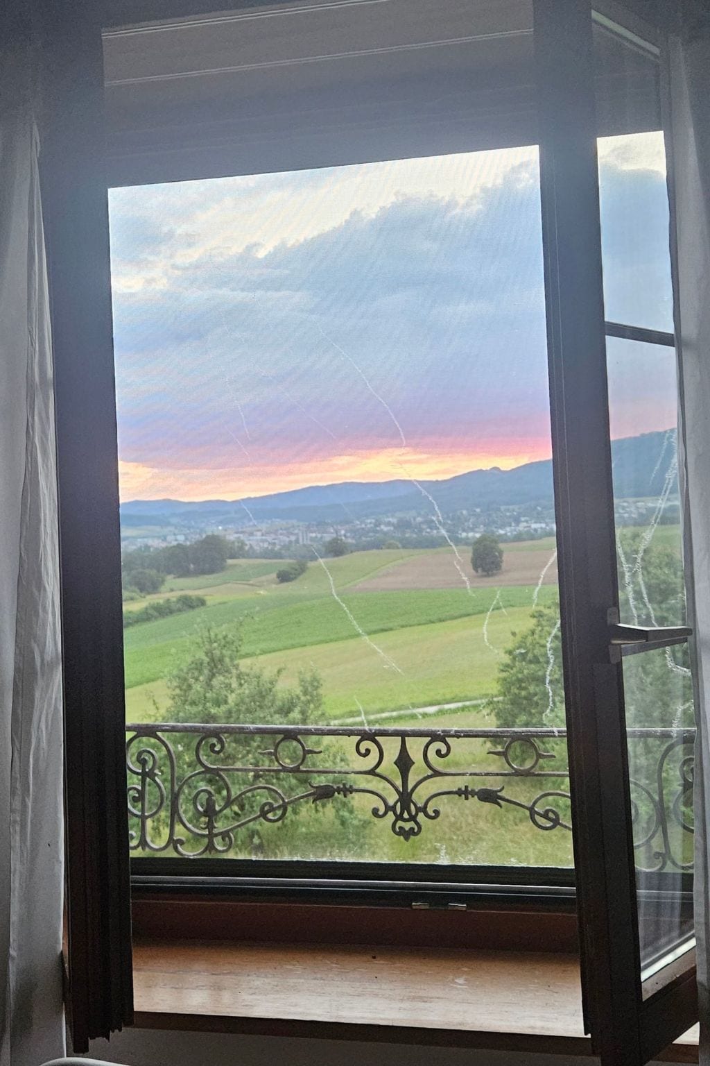 A view through a window with a balcony, featuring a landscape with fields and distant mountains under a cloudy sky.