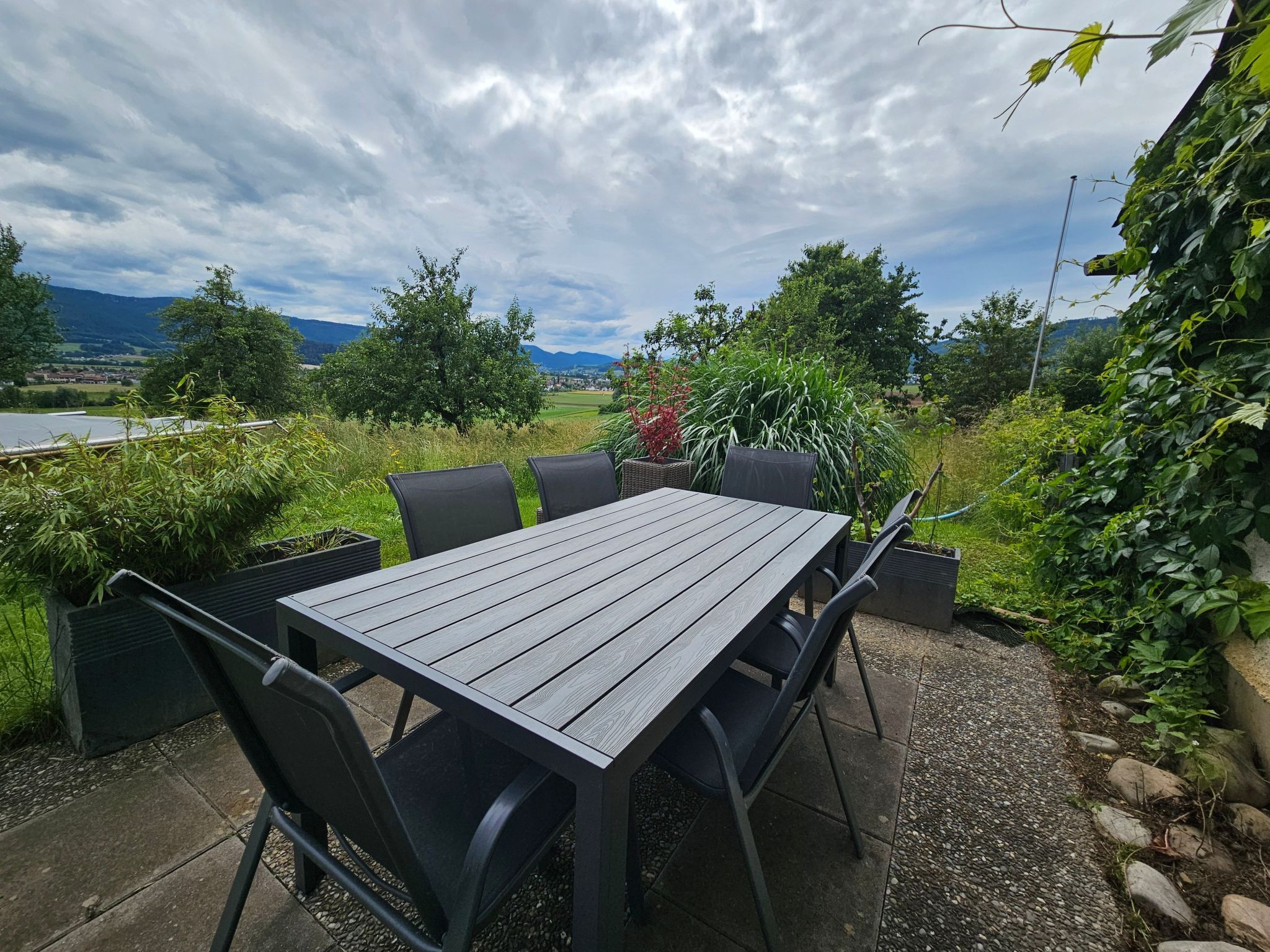 An outdoor patio with a wooden table and chairs, surrounded by greenery and a scenic view.
