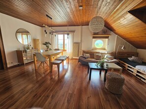 Ein geräumiger Loft mit hohen Decken, Holzbalken, einer Essecke mit rundem Tisch und Stühlen, einer Couch und einem großen Fenster mit Blick.