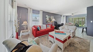 A living room with a red sofa, white armchairs, a white coffee table, and a dining area with a table and chairs. - Stay Where The Rockets Launch & The Waves Roll In! (Satellite Beach)