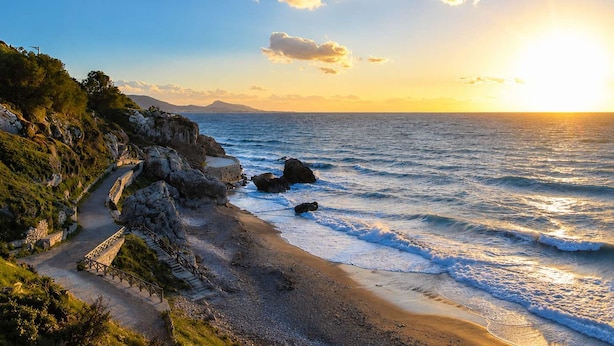 Eine Küstenlandschaft mit einem gepflasterten Weg zum Strand, felsigem Gelände und freiem Blick auf das Meer.