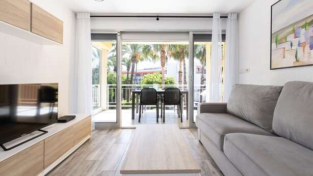 A modern living room with a flat-screen TV, a sofa, a dining table with chairs, and a balcony with a view of palm trees and a hotel building. - C204-058 UHC ANTINEA I APARTAMENTS (Cambrils)