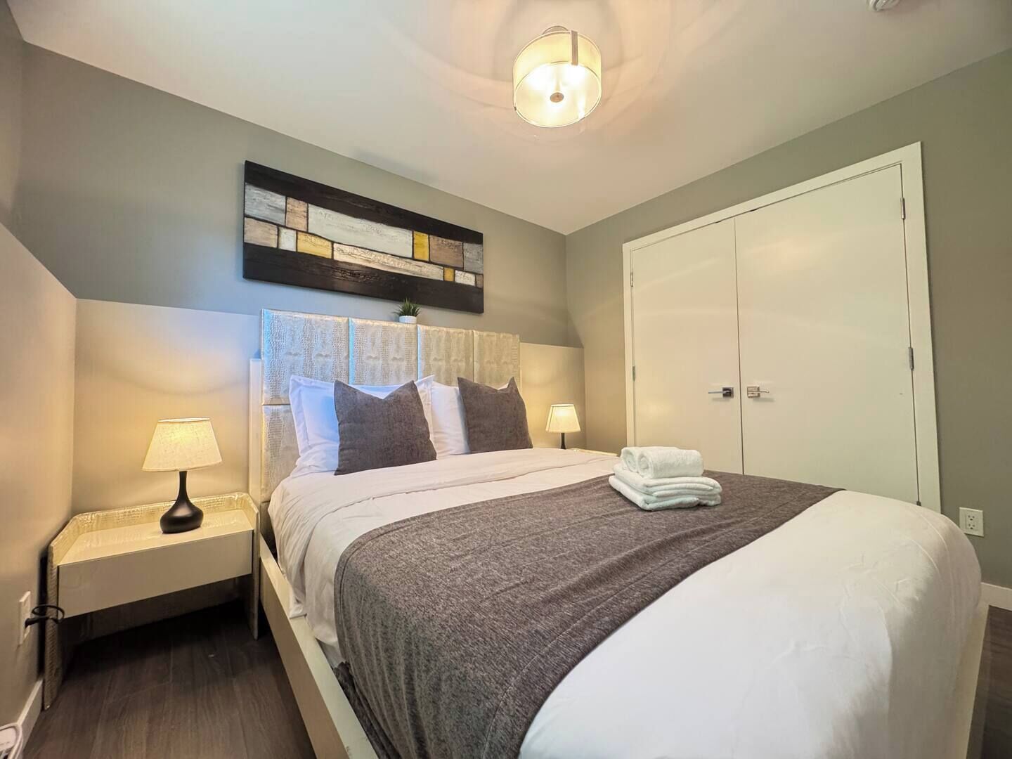 A neatly made bed with pillows, a bedside lamp, a wall art piece, and a wardrobe.