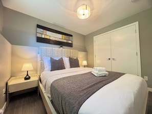 A neatly made bed with pillows, a bedside lamp, a wall art piece, and a wardrobe. - Garden basement suite in Edgemont Village, perfect for Capilano, Grouse Mtn., and FIFA. Quiet, safe area with a bus 5 minutes away for easy downtown access. Walk to shops, cafés, groceries, and more. (North Vancouver)