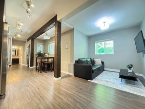A spacious living area with a wooden floor, a dark sofa, a dining table with chairs, and a large window providing natural light. - Garden basement suite in Edgemont Village, perfect for Capilano, Grouse Mtn., and FIFA. Quiet, safe area with a bus 5 minutes away for easy downtown access. Walk to shops, cafés, groceries, and more. (North Vancouver)