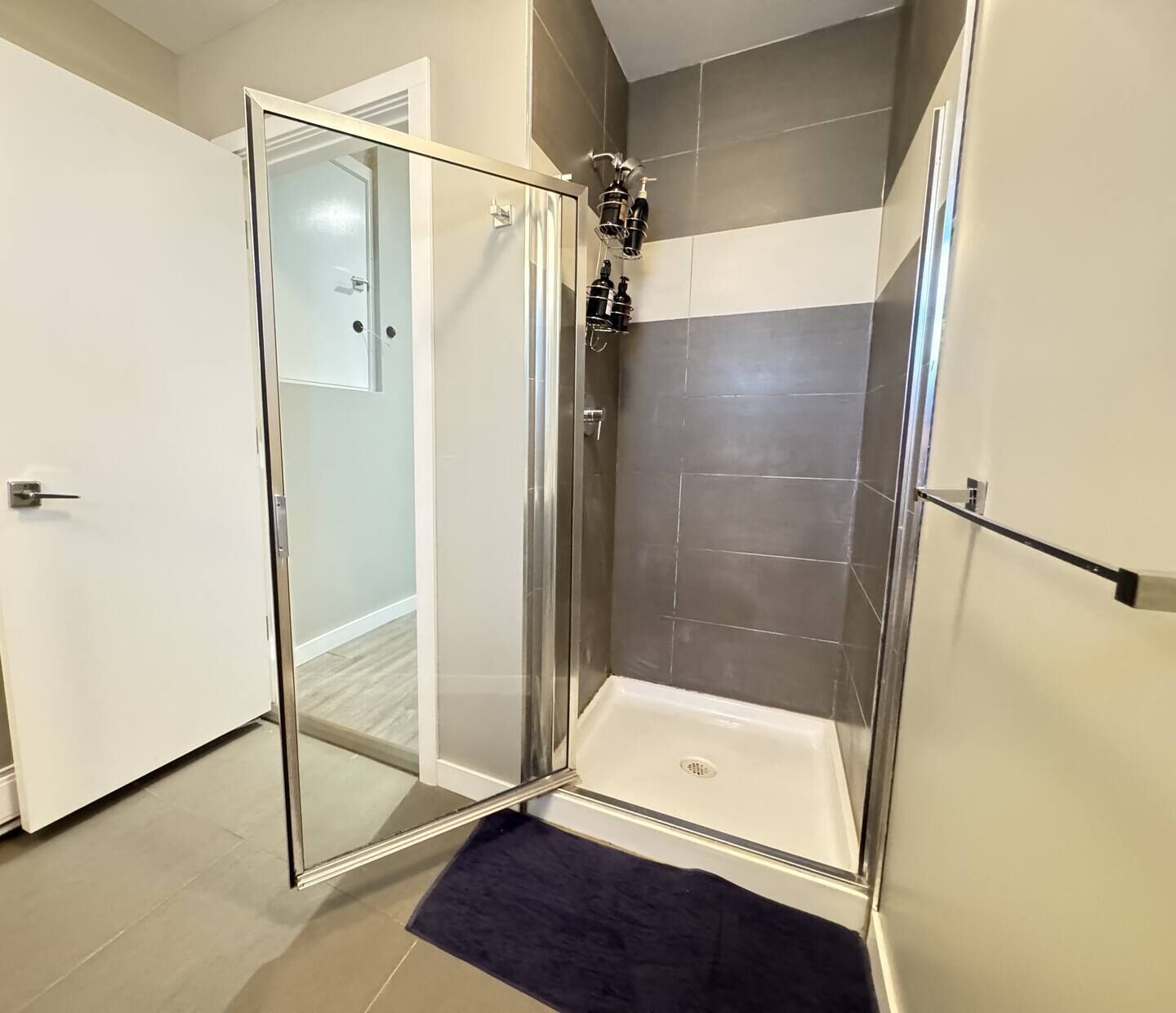A glass-enclosed shower with a white bathtub, grey tiles, and a dark blue mat.