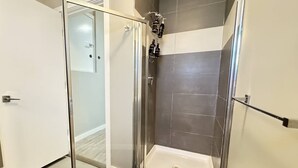 En glasvæg omkranser brusebadet, som har et hvidt badekar, grå fliser og en mørkeblå måtte.