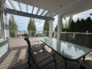 Eine Terrasse im Freien mit einem Glastisch und Stühlen, einem weißen Geländer und Blick auf Bäume und ein Gewässer.