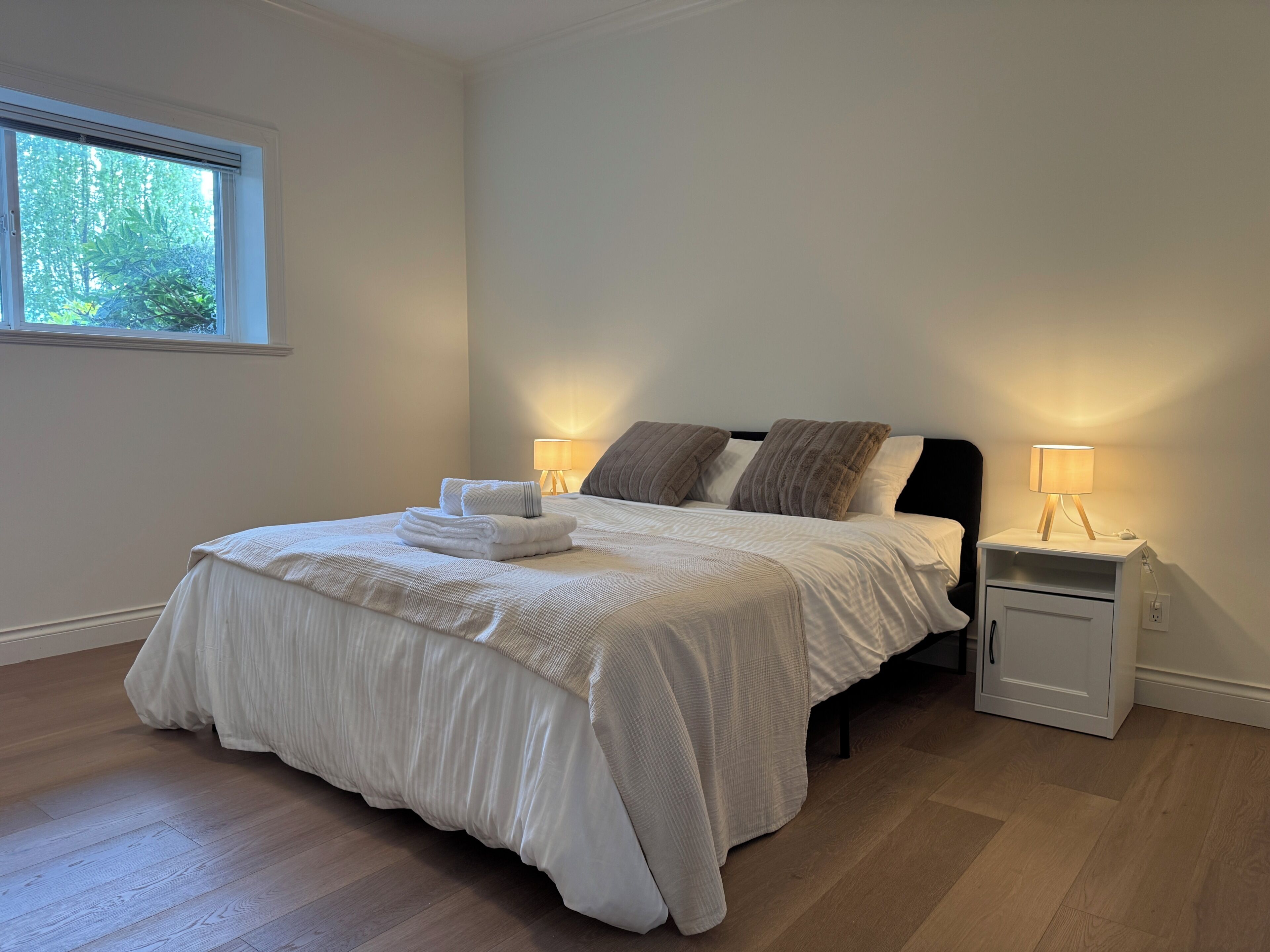 Uma cama bem arrumada com um edredom branco e almofadas, uma mesa de cabeceira com abajur e uma janela com vista para árvores.