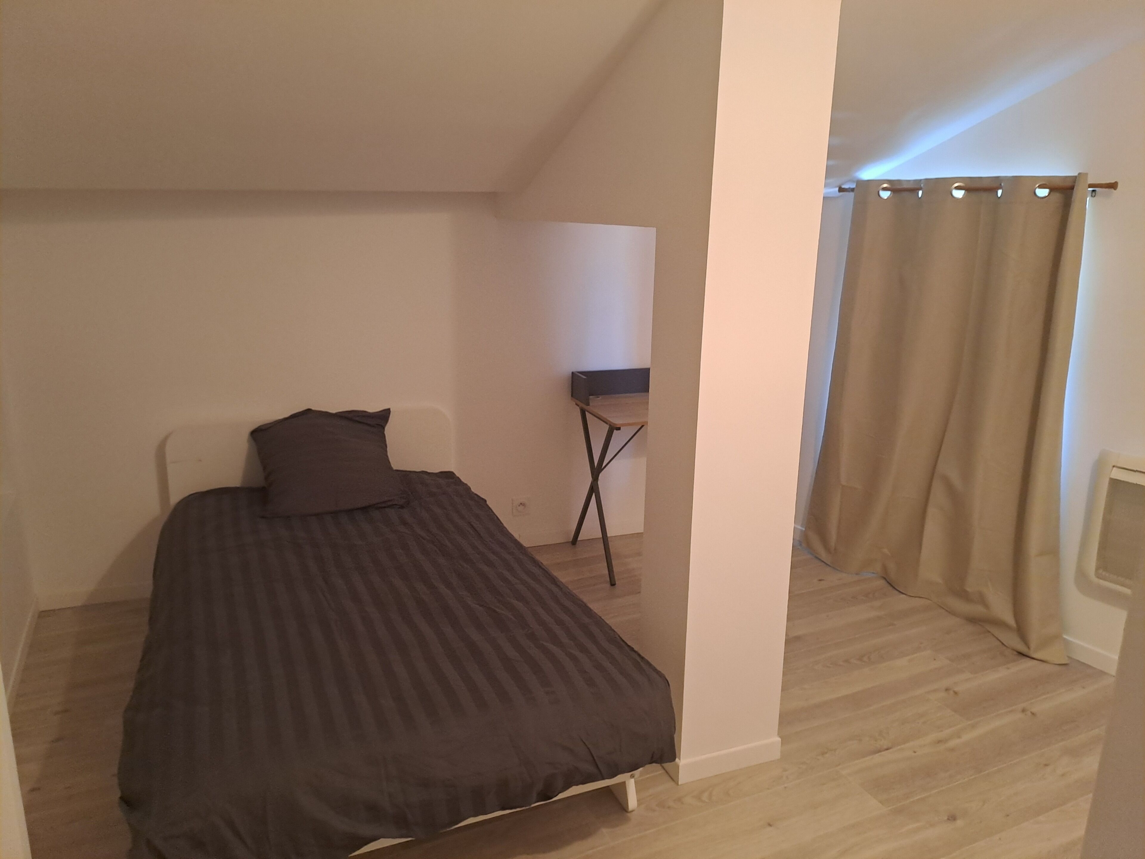 A small, single-bed room with a wooden floor, a bedside table, a lamp, and a curtain.