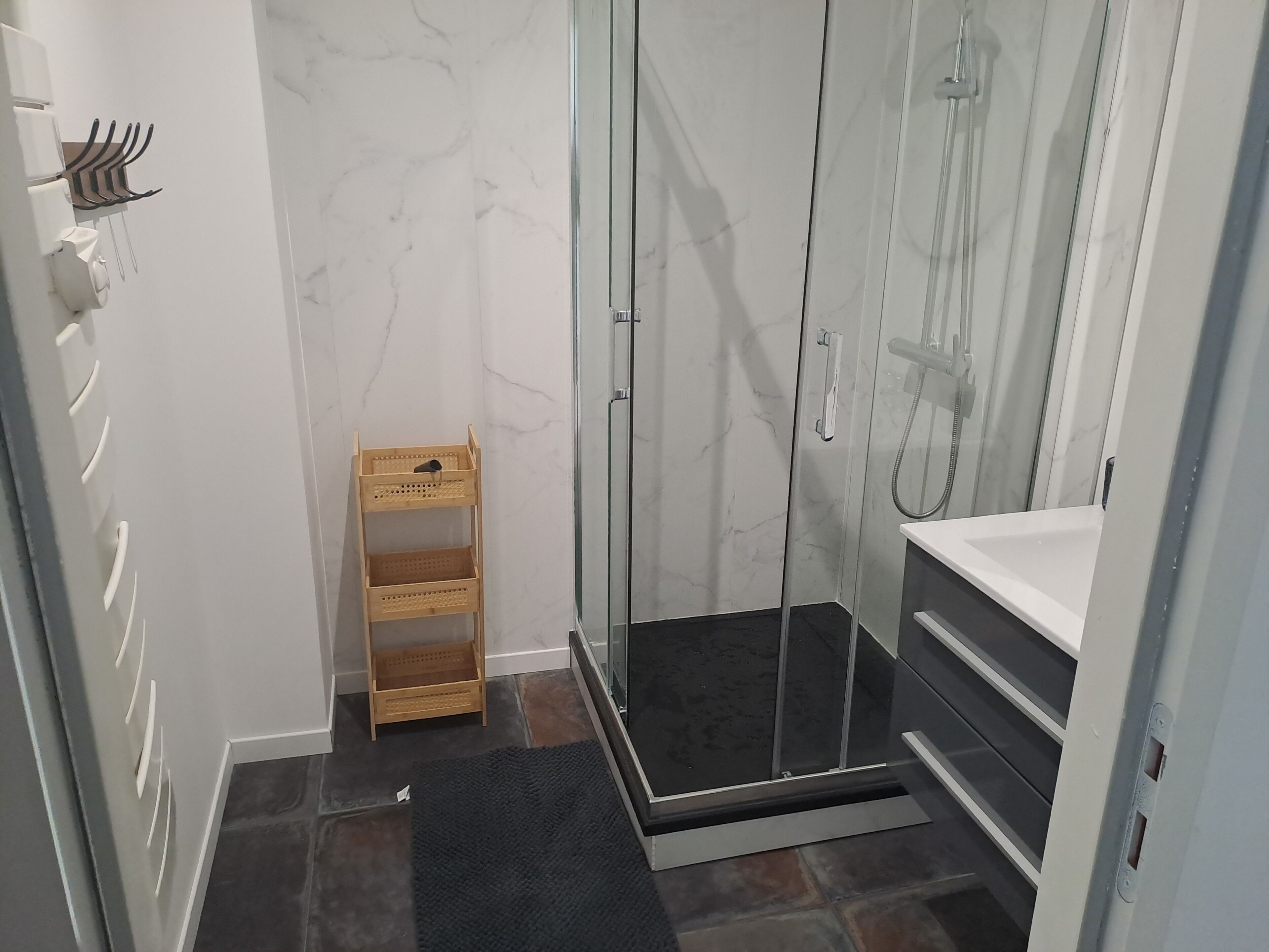 Une salle de bain avec une douche à parois de verre, une étagère en bois et un tapis noir.