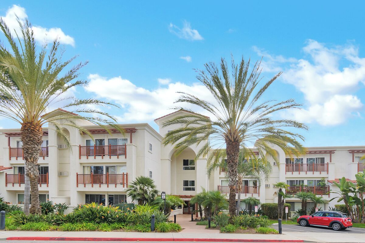 A multi-story hotel with balconies, palm trees, and a red car parked in front.