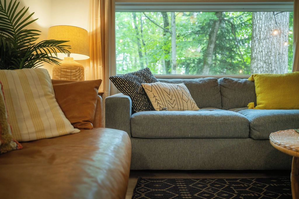 Un salon chaleureux avec un canapé gris, des coussins à motifs, une lampe et une vue sur la verdure à l’extérieur.