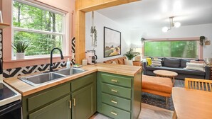 Een moderne keuken met groene kastjes, een houten aanrecht en een spoelbak. Er is een zithoek met een tafel en stoelen, en een bank op de achtergrond.
