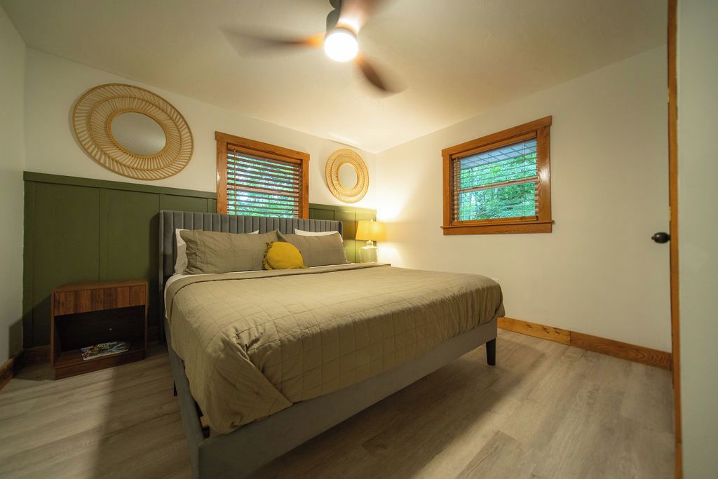 Une chambre à coucher avec un lit, une table de chevet, une lampe et deux fenêtres équipées de stores.