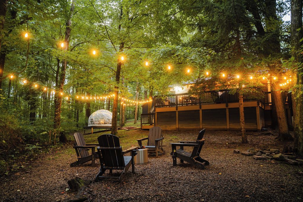 Un espace extérieur aménagé avec des chaises Adirondack, un barbecue et des guirlandes lumineuses, le tout dans un cadre forestier.
