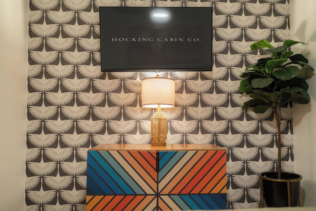 Un téléviseur fixé au mur, avec un papier peint à motifs et une lampe décorative posée sur une table d’appoint.