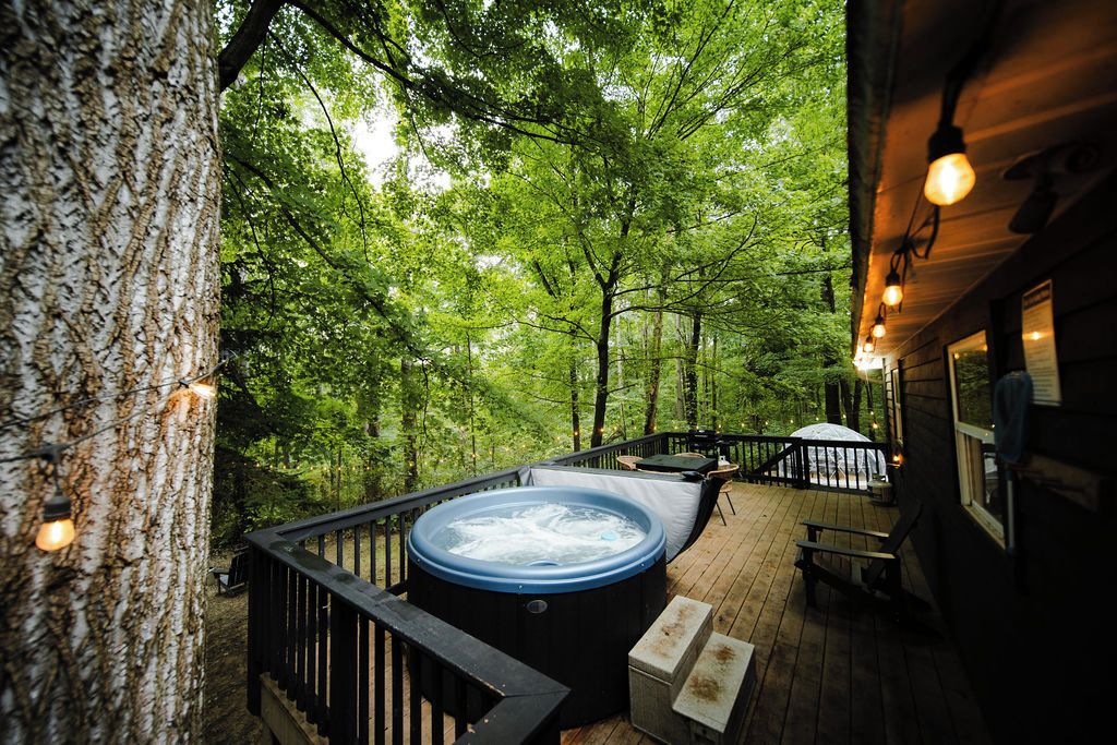 A hot tub on a wooden deck surrounded by trees, with string lights and a bench.