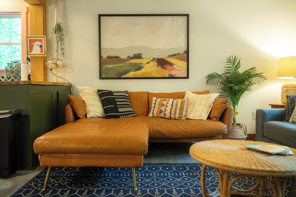 Ein Wohnzimmer mit einem braunen Ledersofa, gemusterten Kissen, einem gerahmten Landschaftsbild, einer Topfpflanze und einem kleinen runden Tisch.