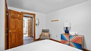Een hotelkamer met een bed, een stoel, een nachtkastje met lamp, een houten deur en een gang naar een andere kamer.