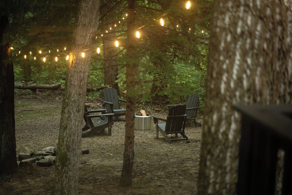 Un espace extérieur aménagé avec des chaises en bois et un foyer, entouré d’arbres et illuminé de guirlandes lumineuses.
