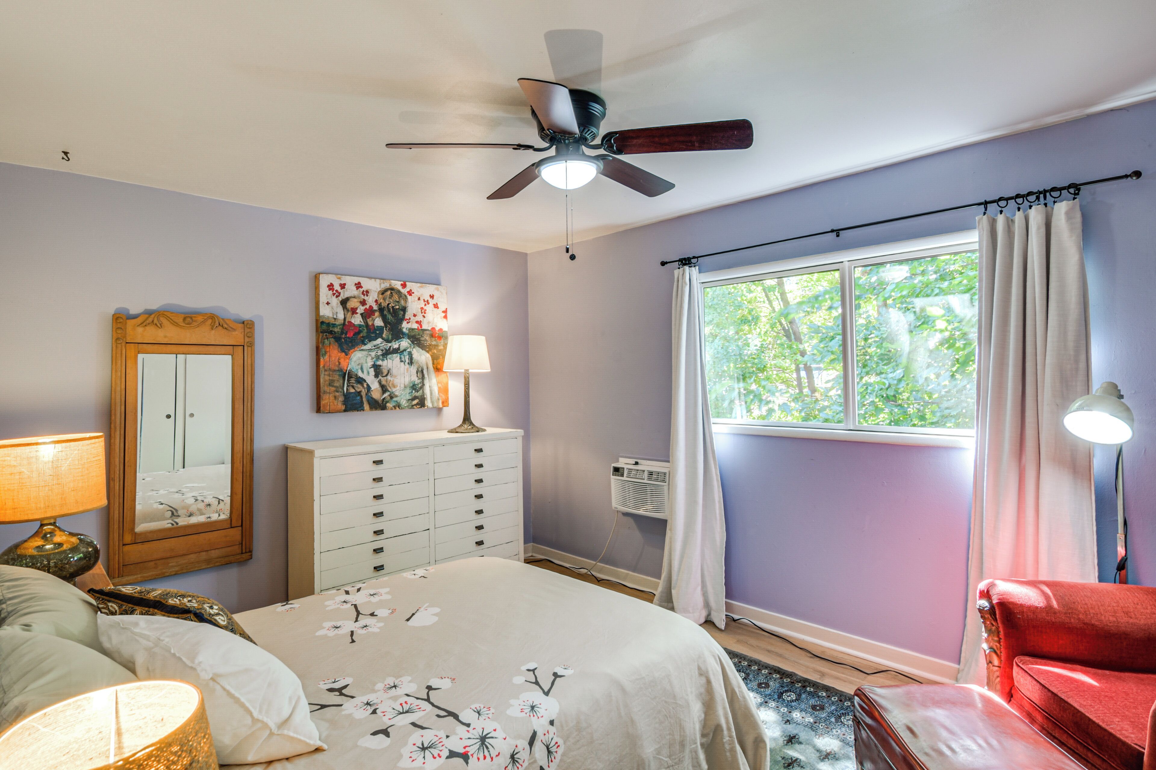 A bedroom with a bed, a dresser, a ceiling fan, a window with curtains, and a red chair.
