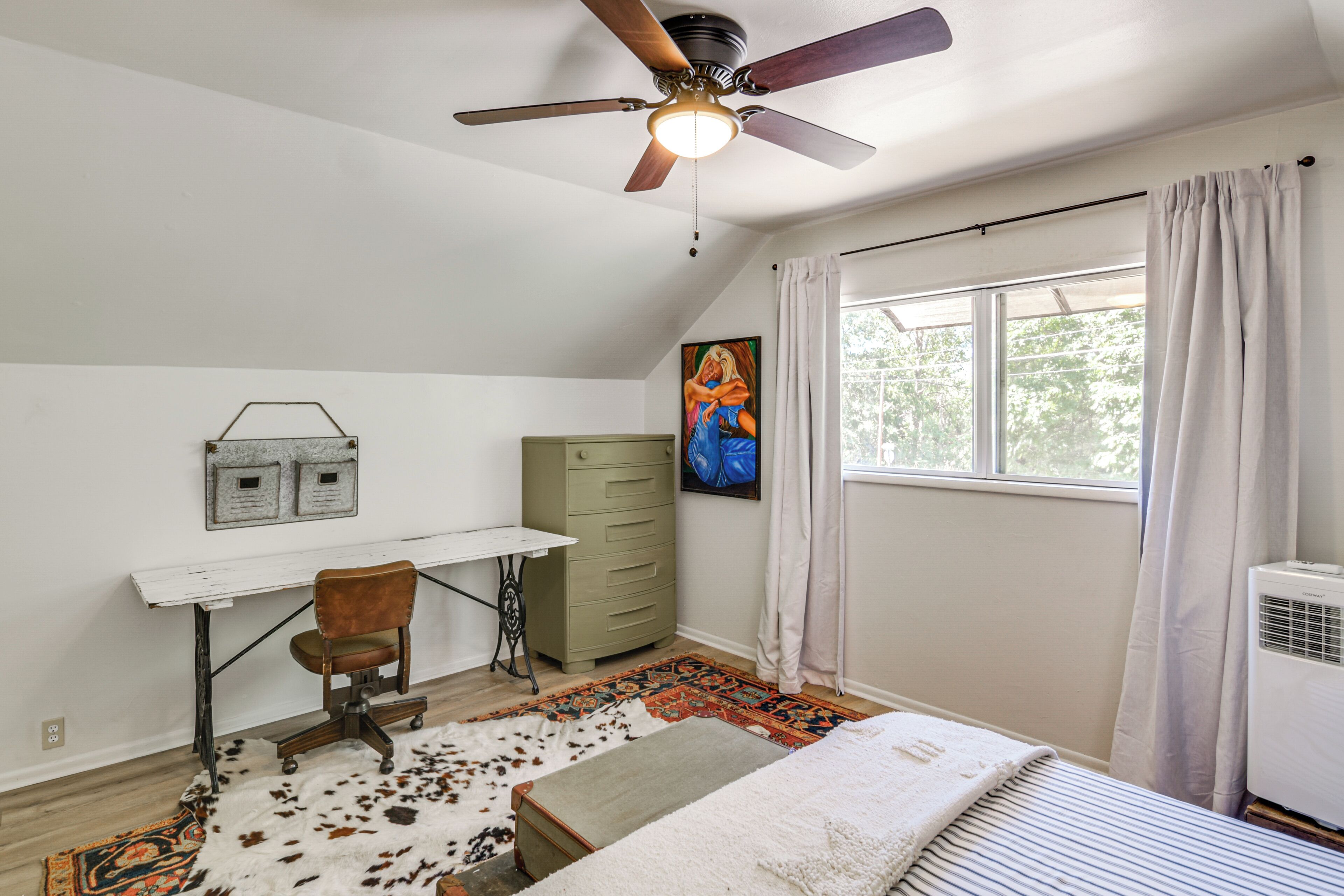 A bedroom with a ceiling fan, a desk, a chair, a bed, a window with curtains, and a radiator.
