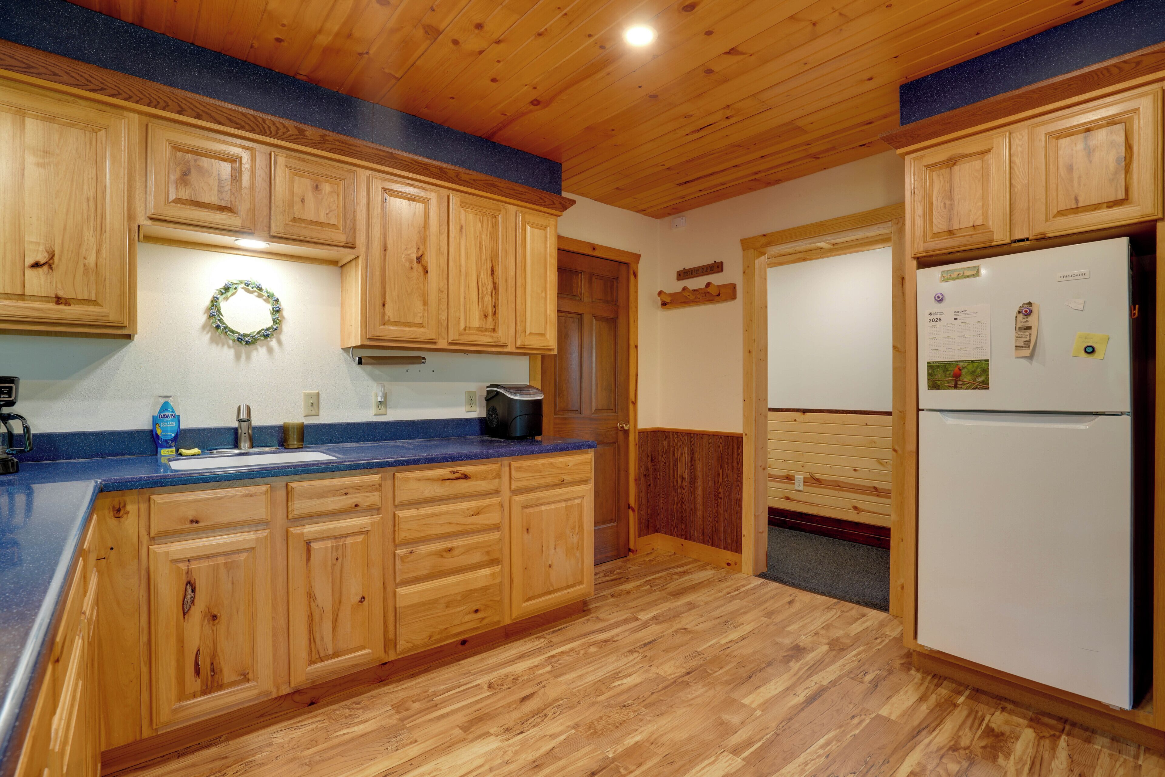 A kitchen with wooden cabinets, a blue countertop, a refrigerator, and a wall-mounted calendar.