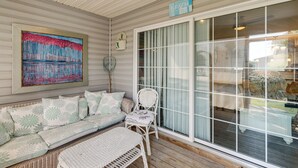 Een overdekt terras met een rieten bank, een witte stoel, en een groot raam met uitzicht op de omgeving.