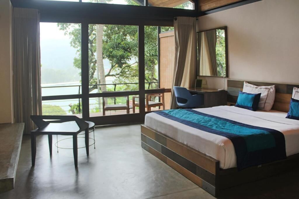 A bedroom with a bed, a desk, and a chair. There is a large window with a view of trees and a body of water.