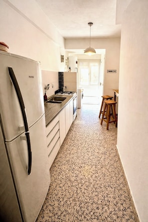 Una cocina estrecha con un refrigerador, un lavamanos y un taburete de madera.