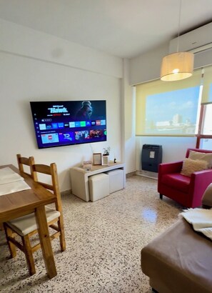 Una sala de estar con una televisión de pantalla plana montada en la pared, una mesa de comedor con sillas, un sofá rojo y una mesita auxiliar con una lámpara.