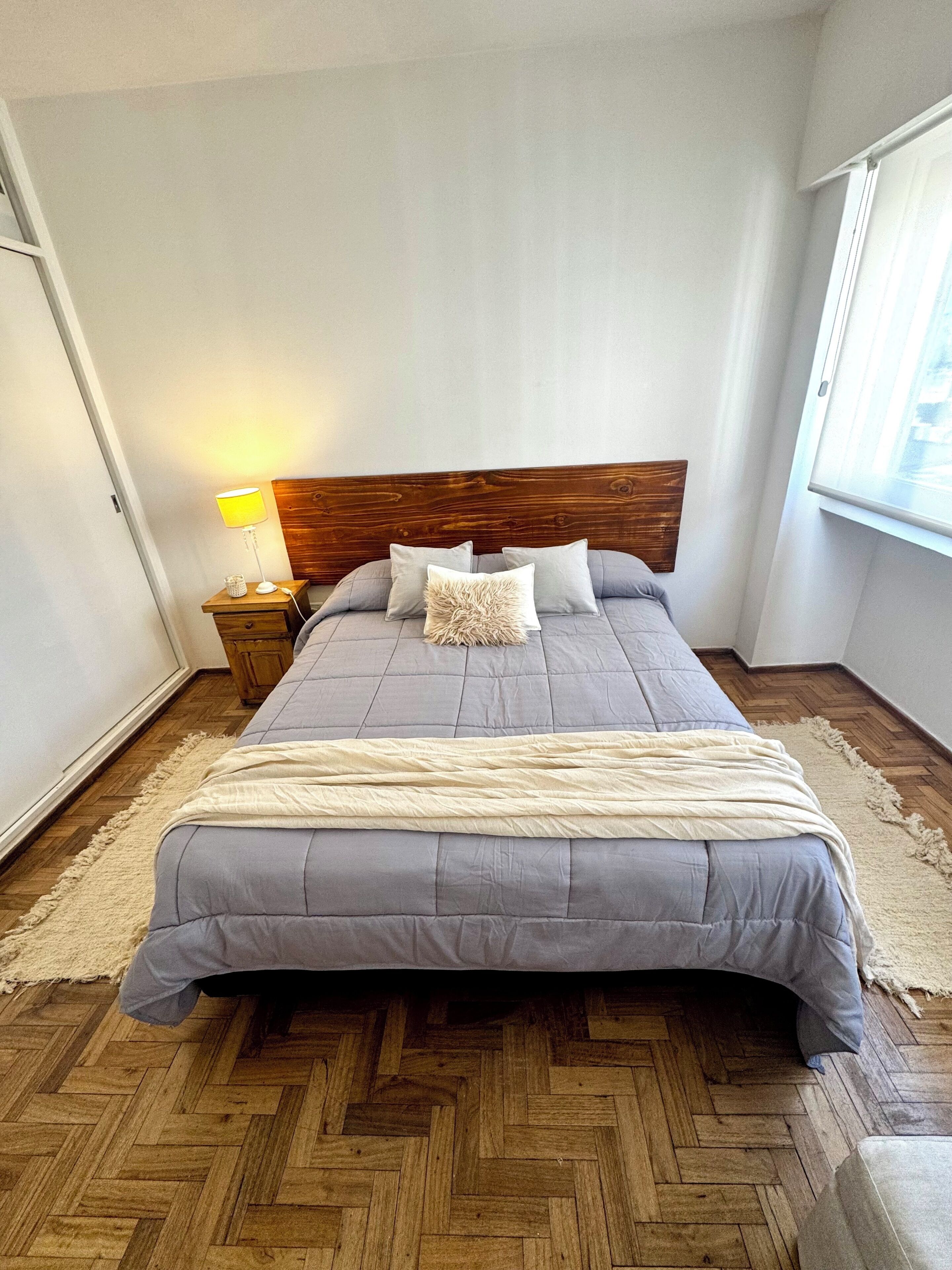 Une chambre à coucher avec une tête de lit en bois, un lit avec une literie grise, une table de chevet avec une lampe et une fenêtre avec des rideaux.