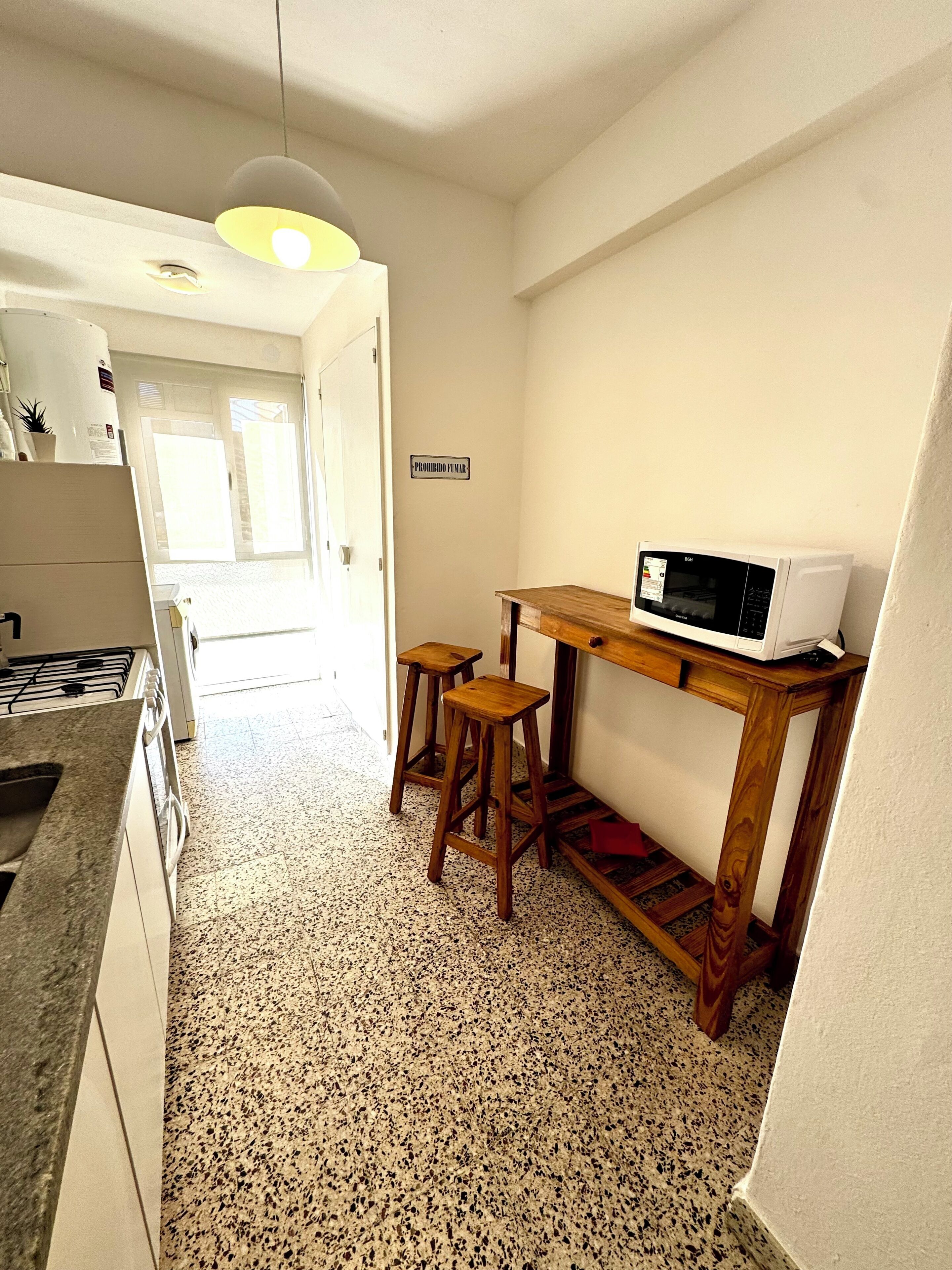 Une petite cuisine avec un four à micro-ondes posé sur une table en bois, deux tabourets, une cuisinière à gaz et un panneau accroché au mur.