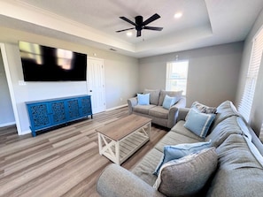 A living room with a flat-screen TV, a ceiling fan, a blue cabinet, and a grey sofa set. - Super Cute 2 bedroom, close to beach, affordable! (Crystal Beach)