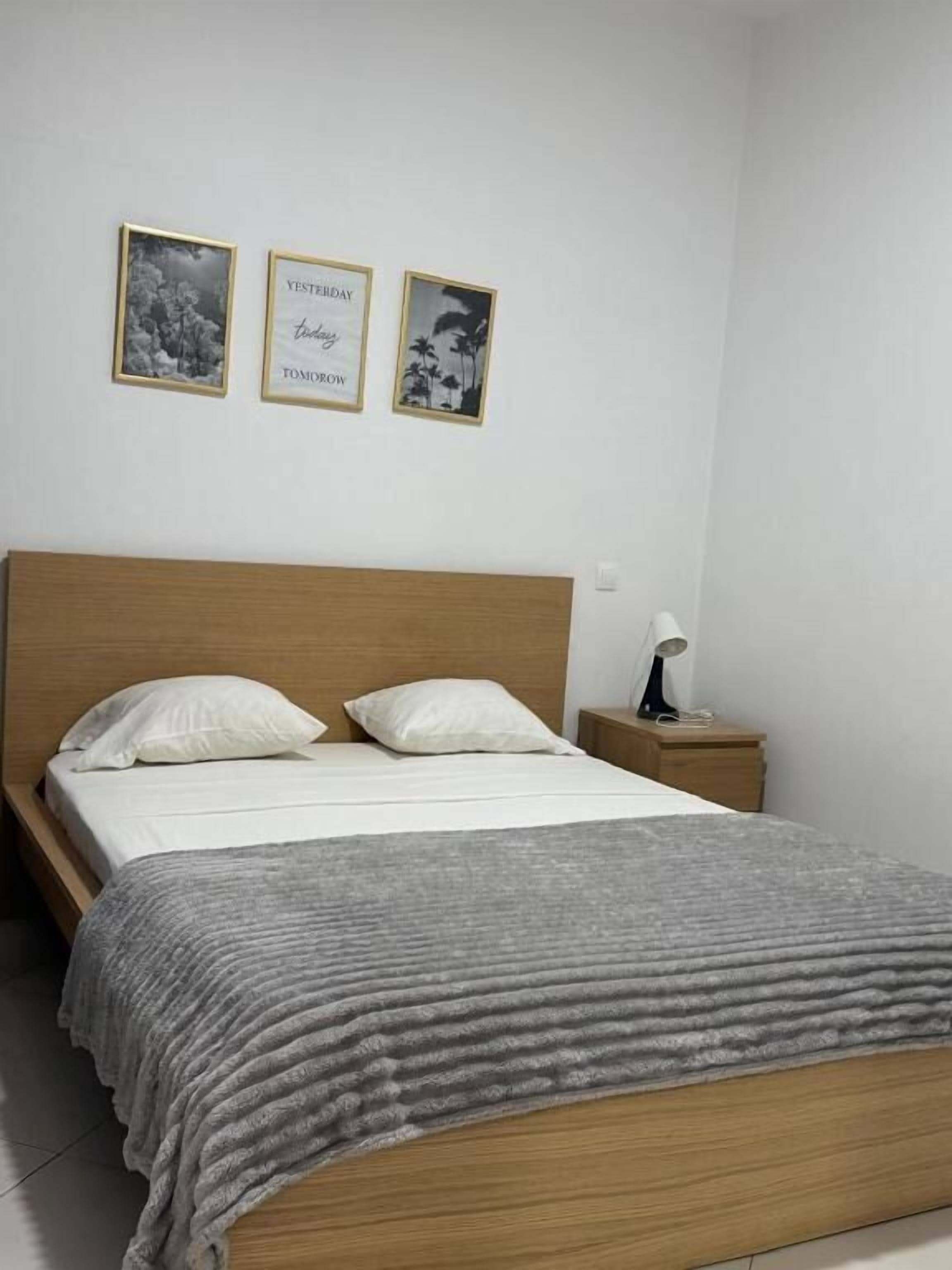 Un lit bien fait, avec une couverture grise, une tête de lit en bois et une table de chevet surmontée d’une lampe. Trois tableaux encadrés sont accrochés au mur au-dessus du lit.