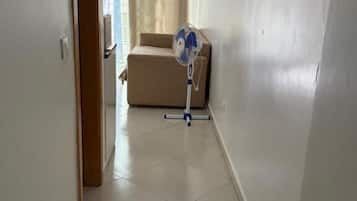 Un couloir avec un canapé beige, un ventilateur sur pied et une porte donnant sur une autre pièce.