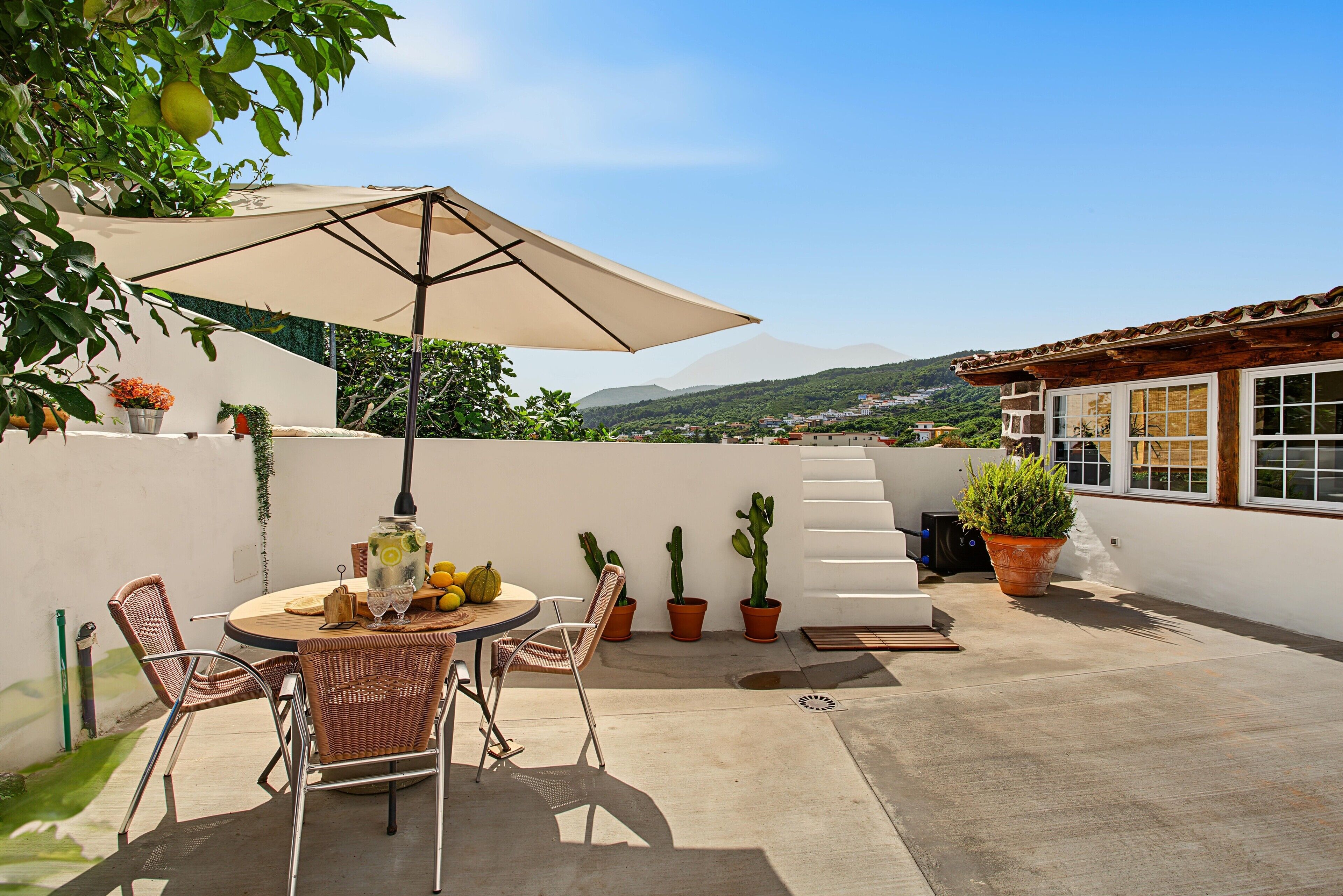 A rooftop terrace with a round table, chairs, an umbrella, potted plants, and a view of a town and mountains.