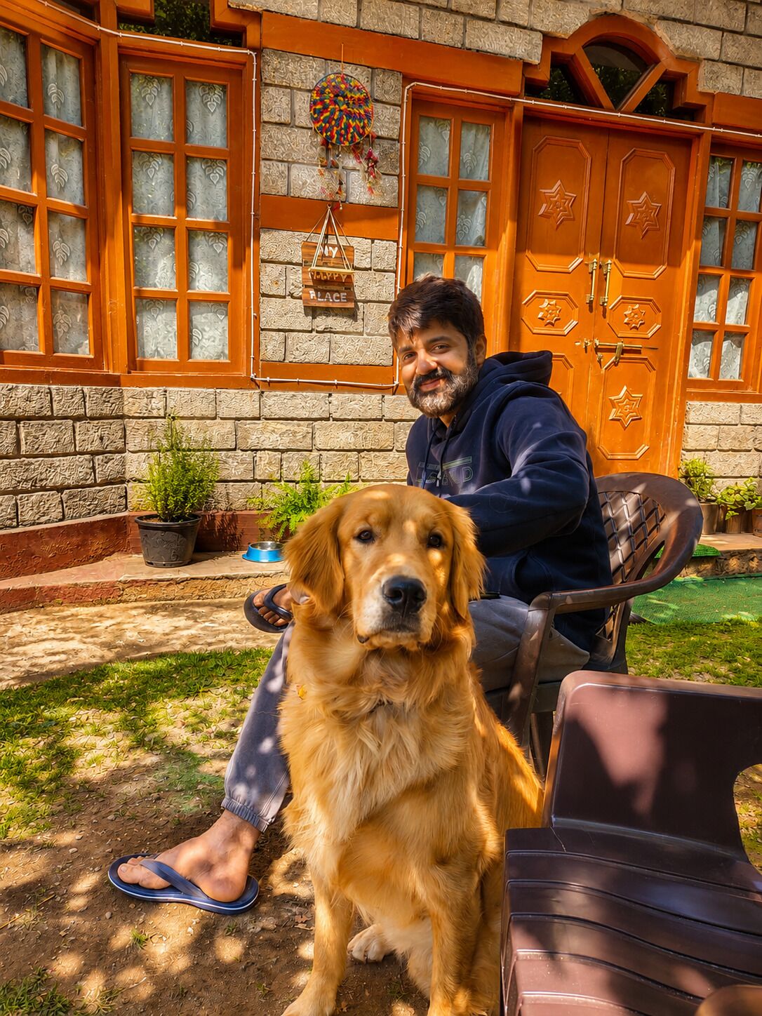 En person sidder i en stol med en hund ved siden af, foran en bygning med en stenfacade og en dør i træ med stjerneformet detaljering.