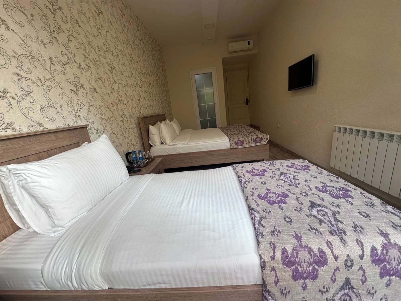 Habitación de hotel con dos camas, un televisor, un radiador y una puerta que da a otra habitación.