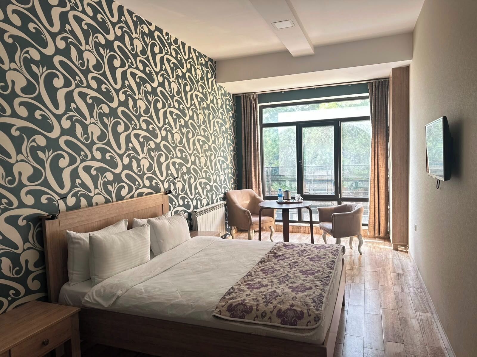 Ein Hotelzimmer mit einem großen Bett, zwei Sessel, einem kleinen Tisch, einem Fernseher und einem Fenster mit Blick auf Grünflächen.