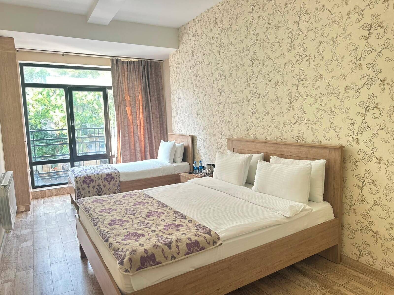 Habitación de hotel con una cama grande, una cama más pequeña, un balcón con cortinas y papel tapiz estampado.